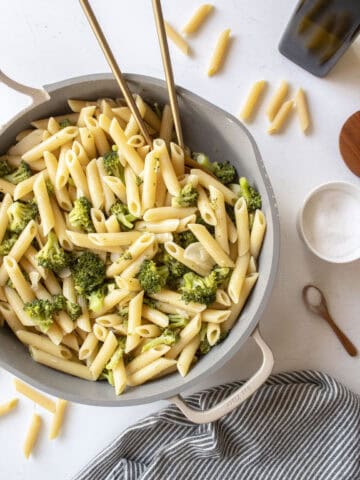 Penne pasta with garlic broccoli in a frying pan with two golden spoons in the pan surrounded by a napkin, salt, and dried pasta.