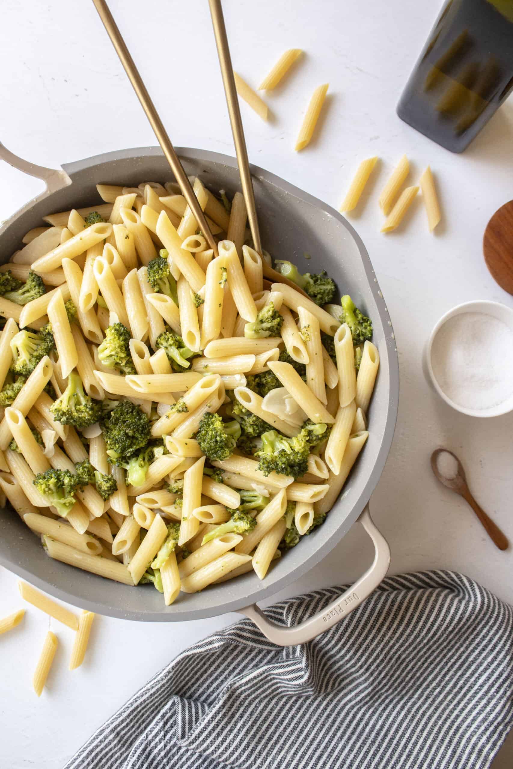 Penne pasta with garlic broccoli in a frying pan with two golden spoons in the pan surrounded by a napkin, salt, and dried pasta.