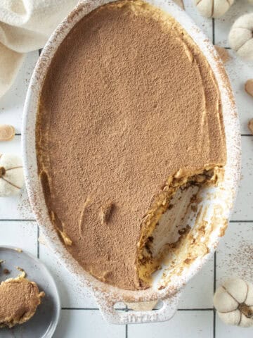 Pumpkin tiramisu with a piece of the tiramisu missing and on a plate to the side surrounded by mini white pumpkins.