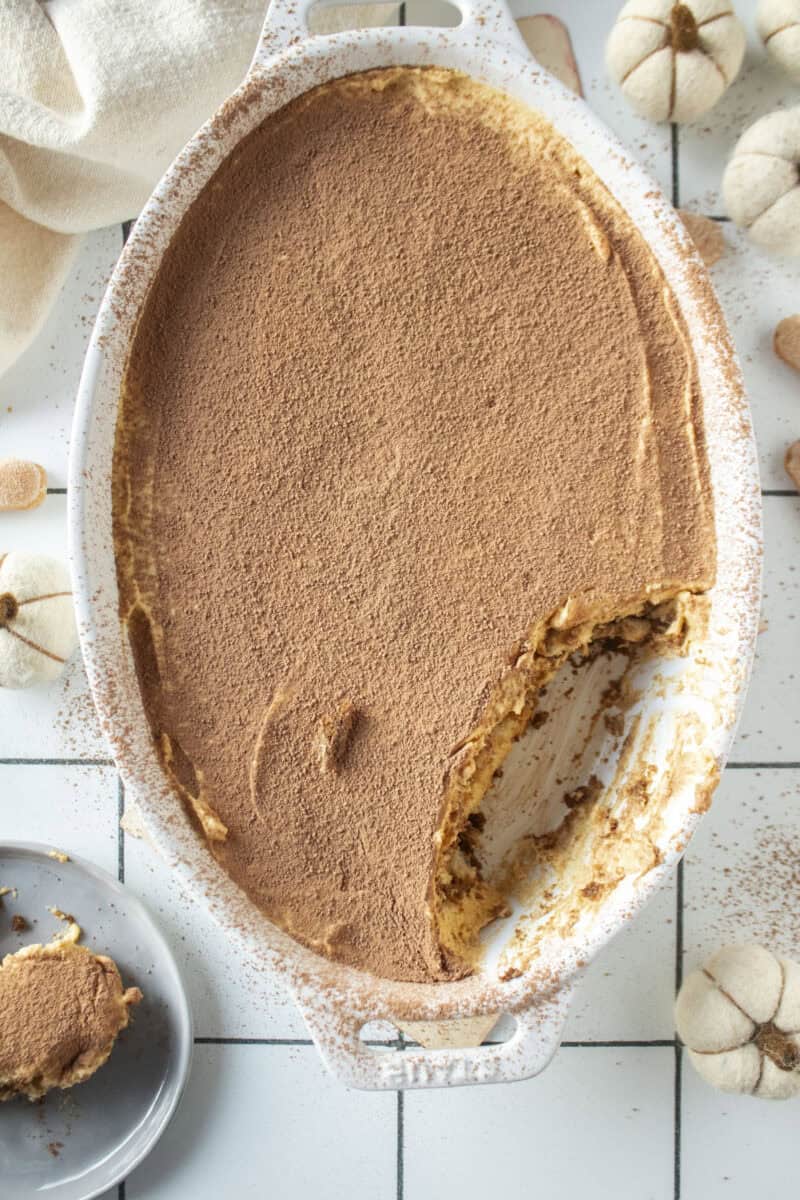 Pumpkin tiramisu with a piece of the tiramisu missing and on a plate to the side surrounded by mini white pumpkins.