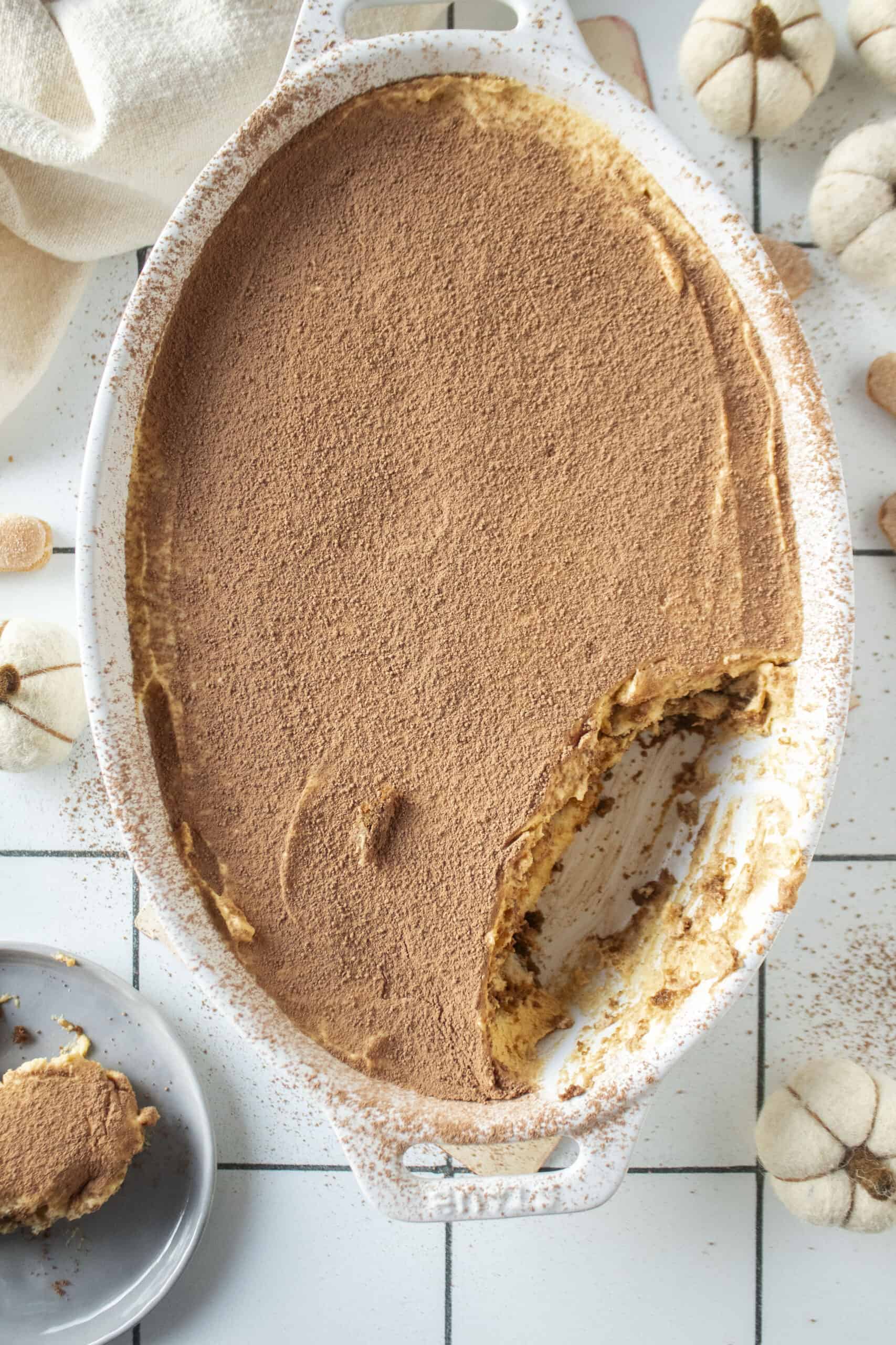 Pumpkin tiramisu with a piece of the tiramisu missing and on a plate to the side surrounded by mini white pumpkins.