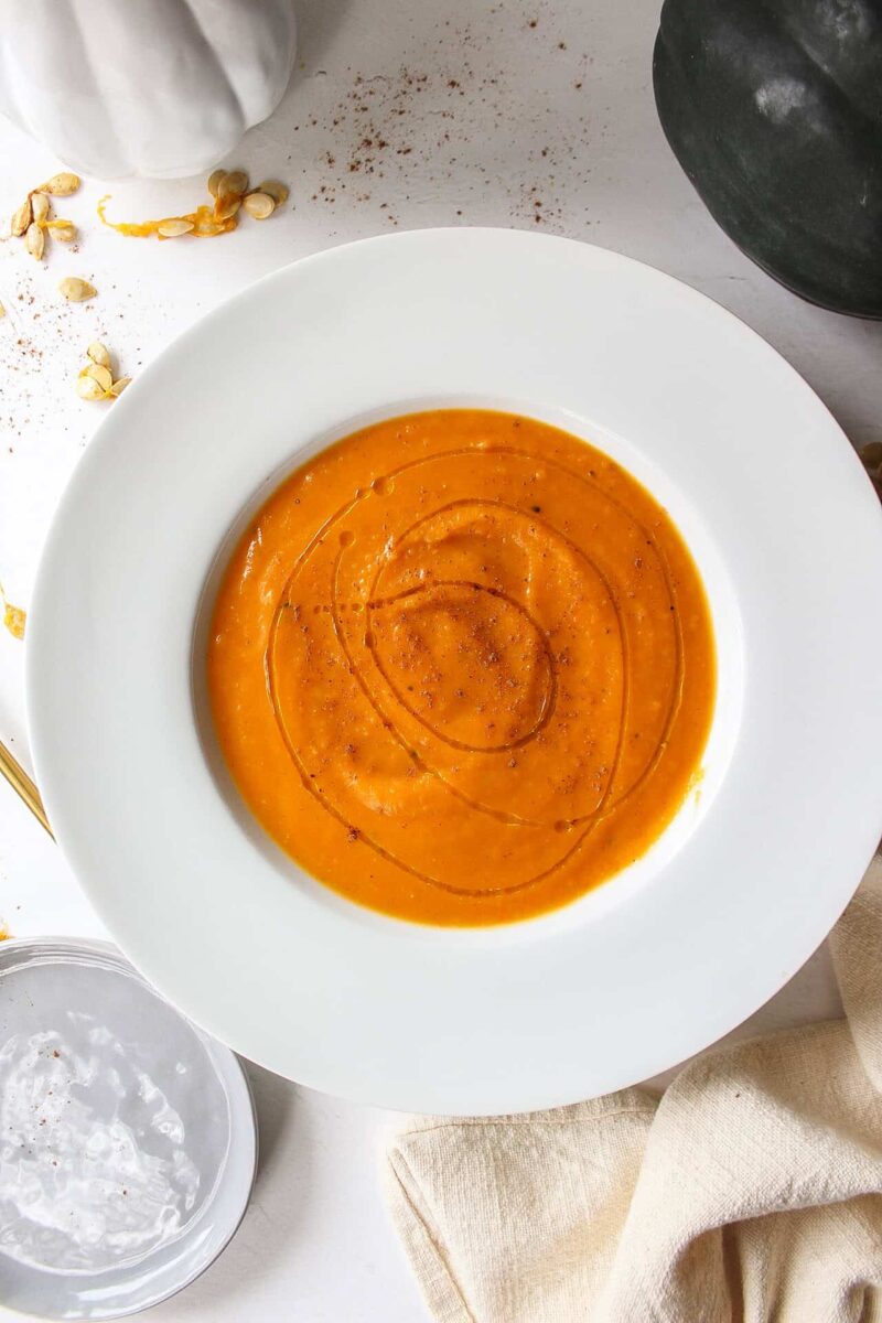 A bowl of acorn squash soup with a drizzle of olive oil on top surrounded by acorn squash and squash seeds.