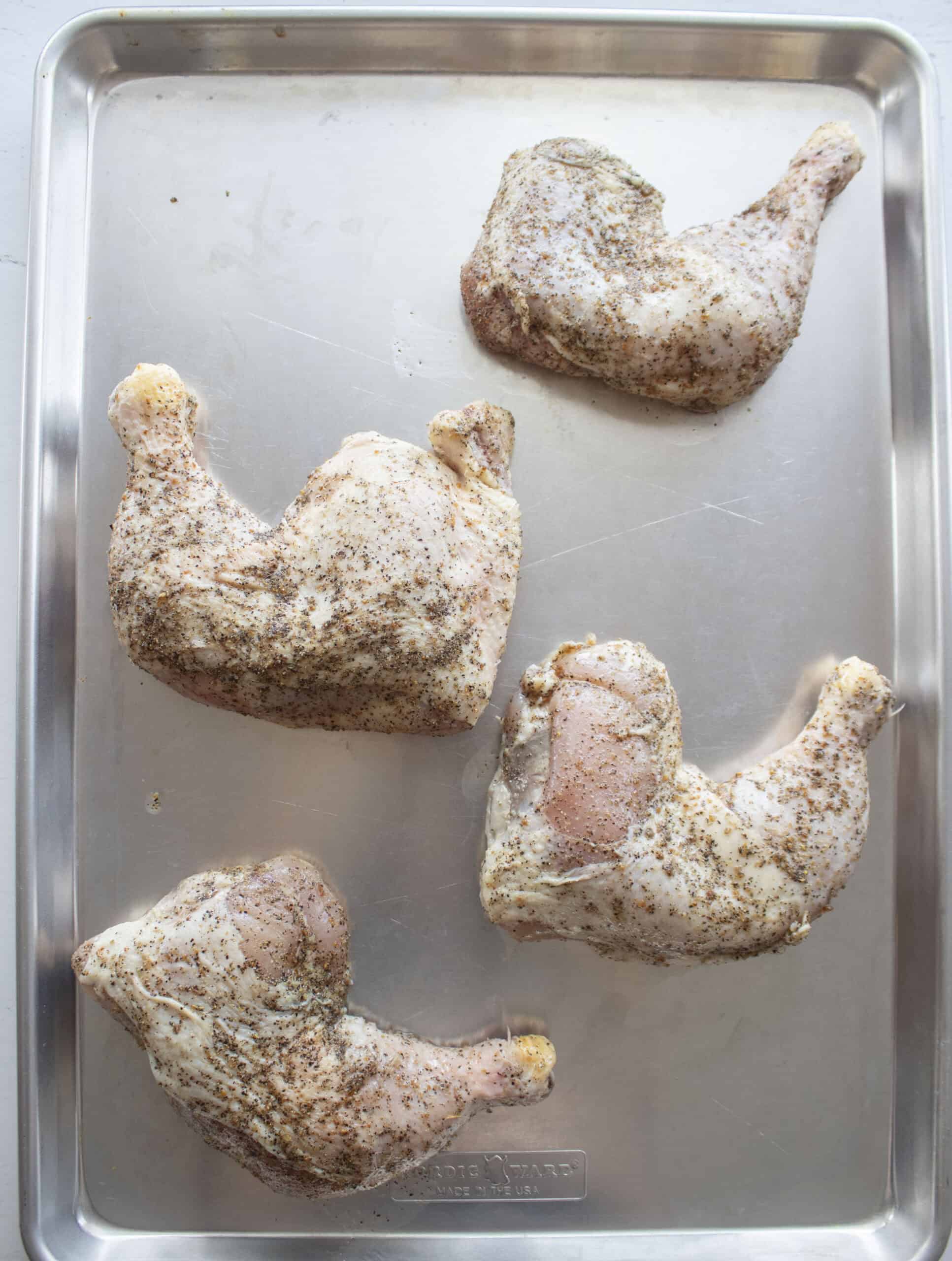 Chicken leg quarters on a baking sheet marinated in lemon pepper seasoning.