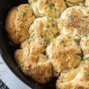 Close up of garlic parmesan crescent roll pull apart rolls baked in a cast iron skillet topped with parsley surrounded by butter and a pastry brush.