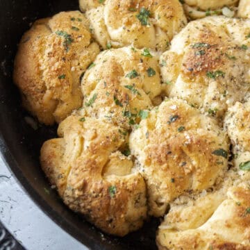 Close up of garlic parmesan crescent roll pull apart rolls baked in a cast iron skillet topped with parsley surrounded by butter and a pastry brush.