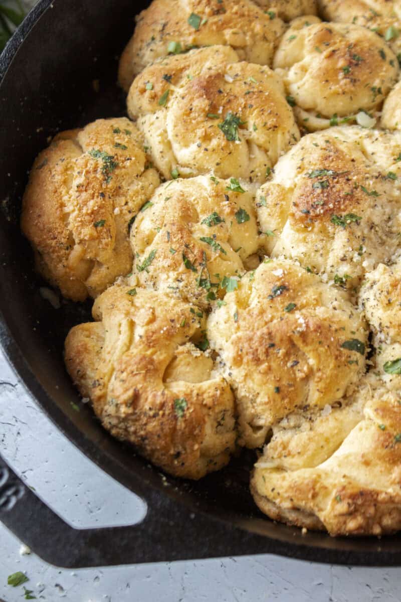Close up of garlic parmesan crescent roll pull apart rolls baked in a cast iron skillet topped with parsley surrounded by butter and a pastry brush.