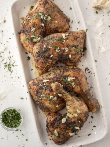 Four baked lemon pepper chicken leg quarters topped with a melted garlic butter with parsley on a serving platter.
