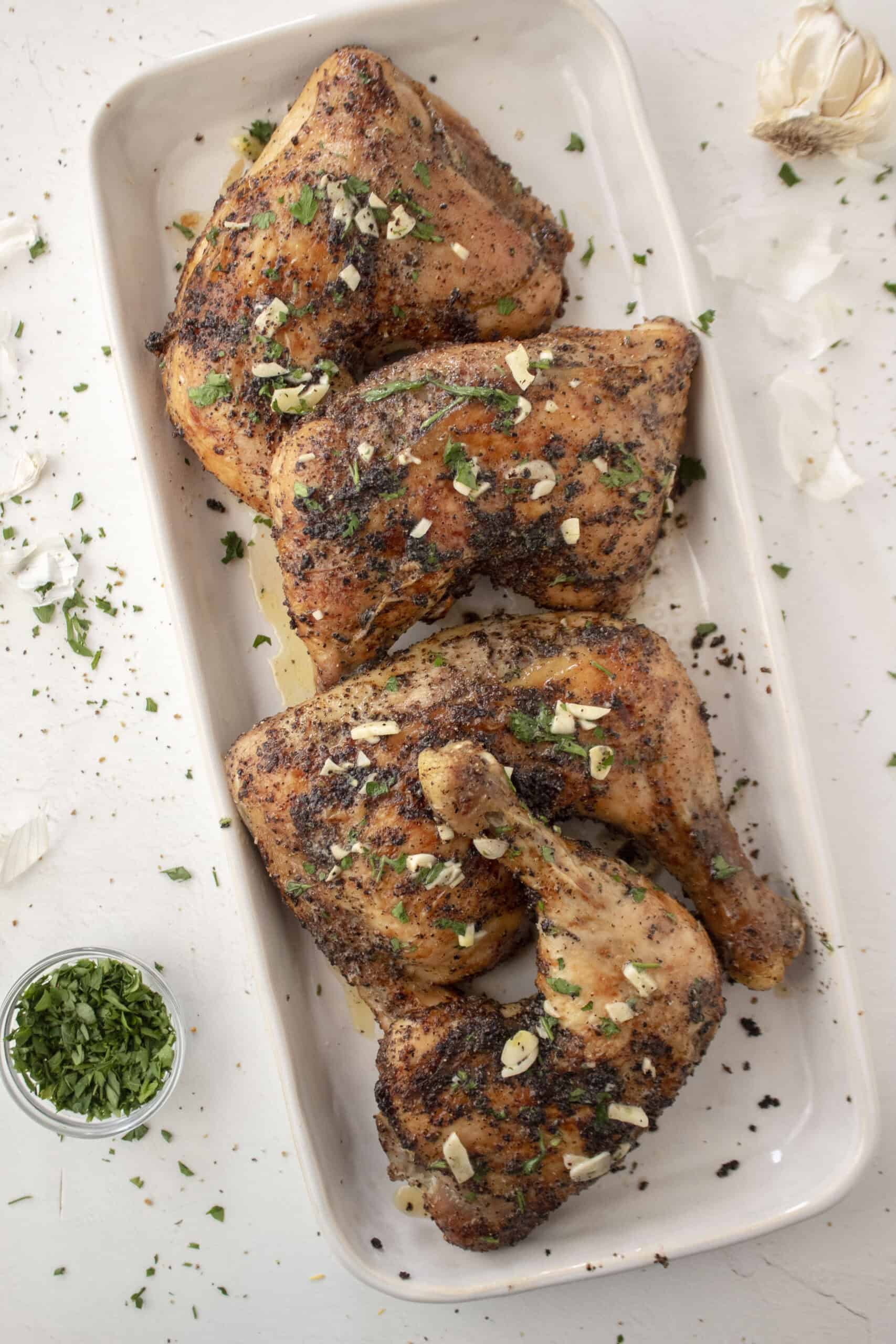 Four baked lemon pepper chicken leg quarters topped with a melted garlic butter with parsley on a serving platter.