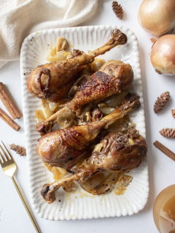 Four apple cider braised turkey drumsticks on a serving platter with cooked onions.