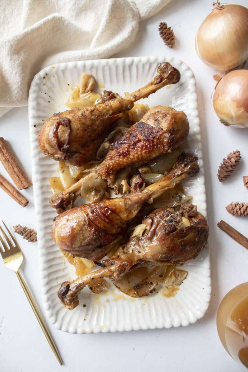 Four apple cider braised turkey drumsticks on a serving platter with cooked onions.