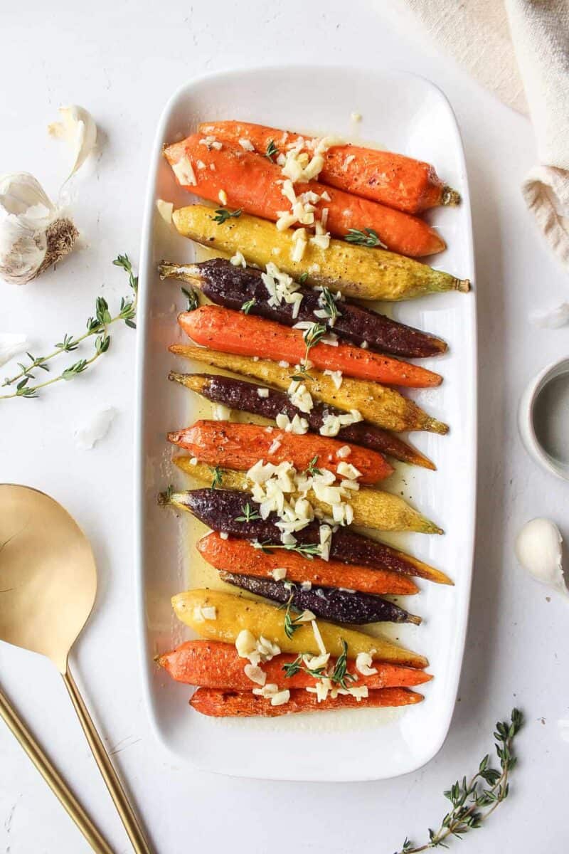 Garlic butter rainbow carrots on a white serving platter surrounded by gold serving utensils and thyme.