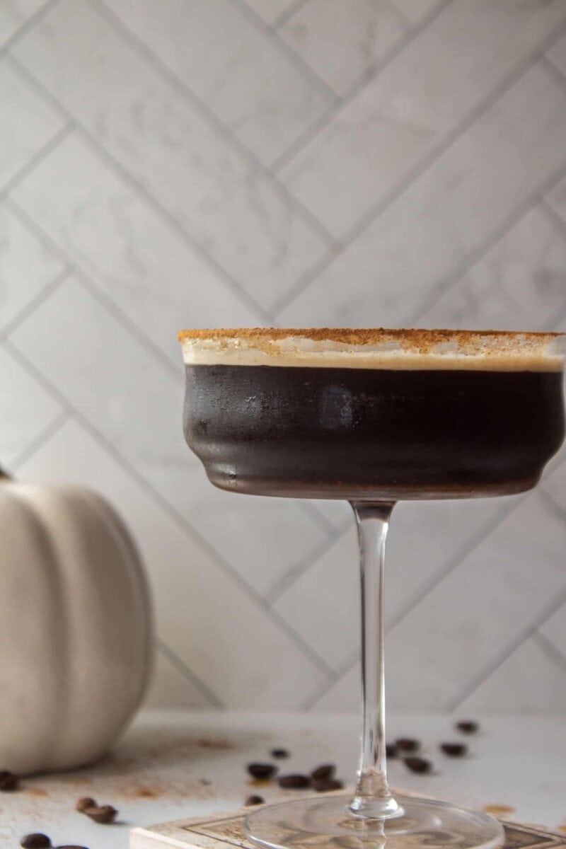 A pumpkin spice espresso martini in a coupe martini glass next to a white porcelain pumpkin